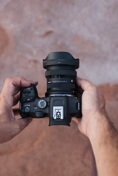 Photographer's hands holding a mirrorless camera with wide-angle lens attached, viewed from above against a textured brown surface.
