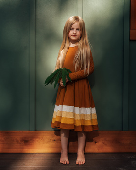Young girl with long reddish-blonde hair wearing an orange and white striped dress, standing barefoot against a green panel wall.
