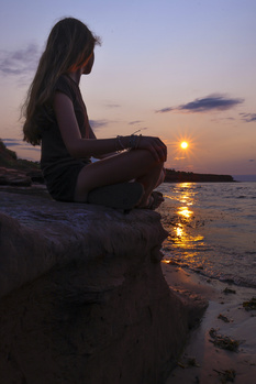 Woman sitting on a rock by the ocean during golden hour sunset with calm water reflecting warm light.