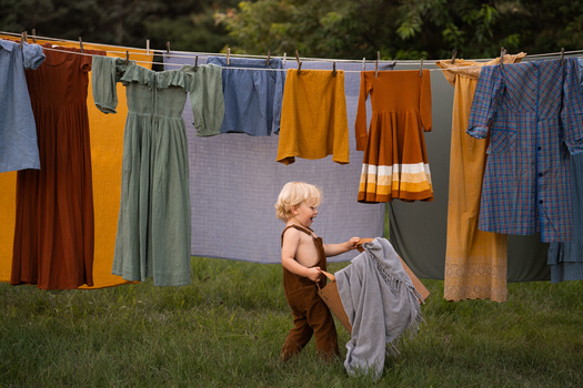 Toddler holding wet laundry on grass beneath clothesline with colorful garments drying in sunlight.