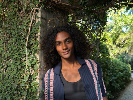 Woman with curly dark hair smiling at camera while leaning against ivy-covered wall in dappled sunlight.