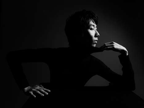 Profile portrait of a person in dark clothing against a black background, lit with dramatic side lighting.