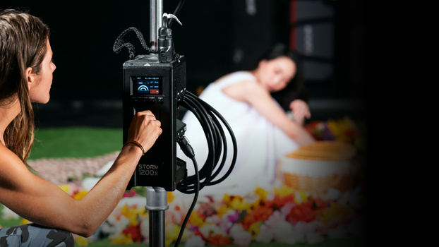 Photographer operating a monolight on a stand during an outdoor photoshoot with a model in the background.