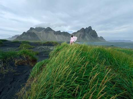 Person in white shirt standing on grassy volcanic terrain with dramatic dark mountains in the background.