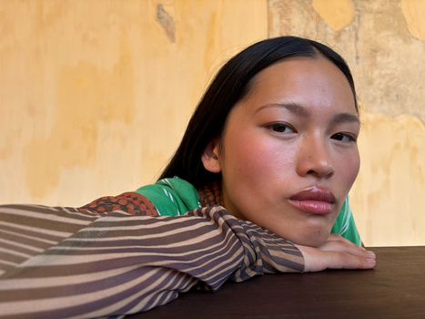Portrait of a woman with dark hair resting her arm on a wooden surface, wearing striped and green clothing against a textured yellow wall.