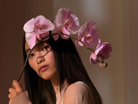 Woman with long dark hair wearing a pink orchid flower crown against a warm neutral background.
