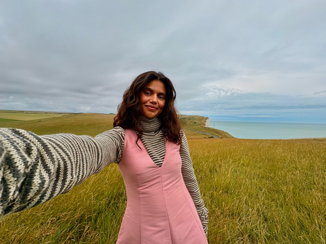 Woman in pink vest and striped shirt smiling at camera in vast coastal grassland.