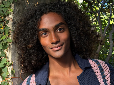 Woman with voluminous curly hair smiling at camera against tree trunk and ivy backdrop.