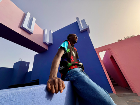 Person sitting on blue beam against colorful geometric architecture with pink and blue structures.