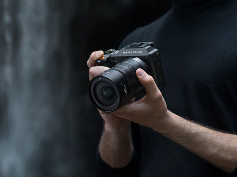 Photographer holding a medium format camera with a lens attached, shot against a blurred gray stone background.