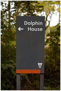 Directional wayfinding sign with dark gray background pointing left to Dolphin House, set against blurred green foliage.