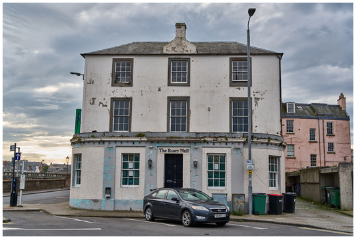 White corner pub building with blue storefront beneath dramatic cloudy sky and parked car.