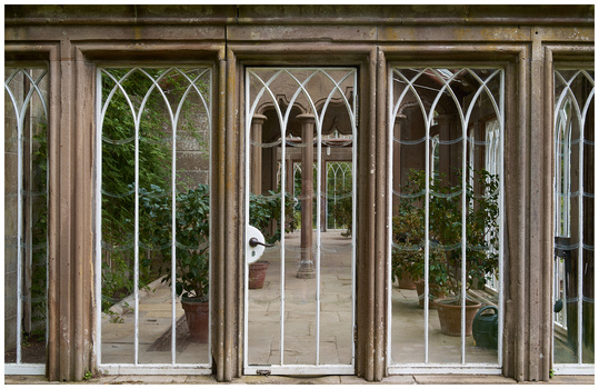 View through Gothic-style glazed doors into a sunlit courtyard with potted plants and stone flooring.