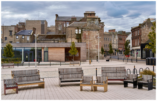 Public plaza with contemporary benches and seating facing historic stone buildings under cloudy sky.