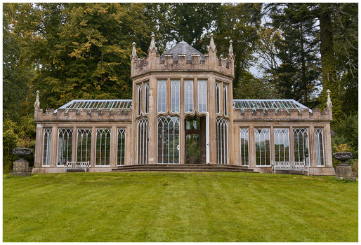 Ornate stone garden pavilion with turrets, arched windows, and pitched roofs flanked by manicured lawn and trees.
