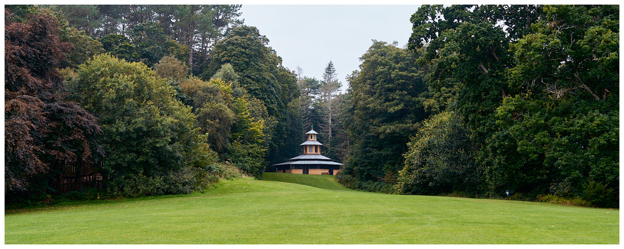 Symmetrical landscape with a small domed structure centered in an expansive green field, flanked by dense forest.