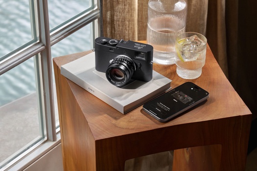 Black rangefinder camera on white pedestal beside smartphone and water glasses on wooden table by window.