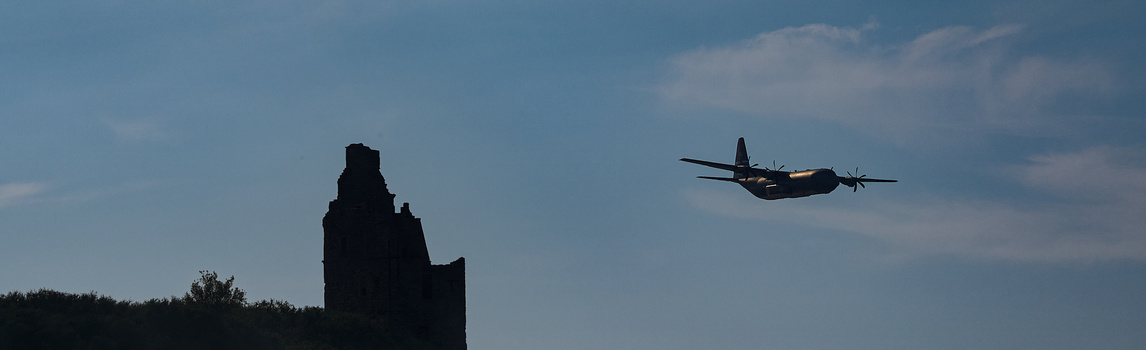 Military transport aircraft flying past a silhouetted ruined tower structure against a blue sky.
