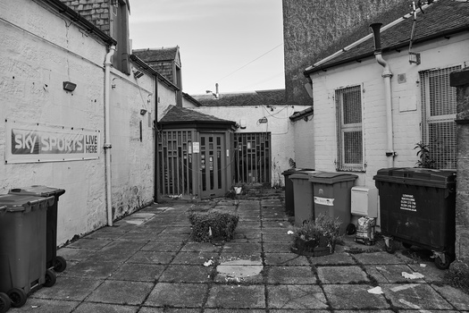 Narrow residential alleyway with white-painted brick buildings, dark garbage bins, and wet pavement in black and white.