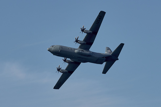 Military transport aircraft with four propellers photographed from below against a clear blue sky.