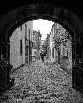 Cobblestone street framed by a dark stone archway, with historic buildings and pedestrians visible down the narrowing alley.