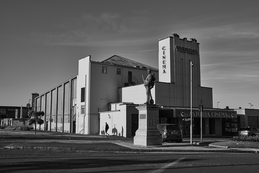 Black and white photograph of a modernist cinema building with vertical tower and statue in foreground.