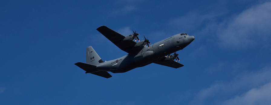 Military transport aircraft in flight against a clear blue sky, viewed from below with twin propellers visible.