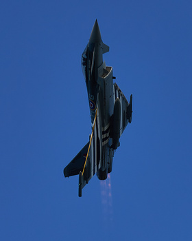 Military fighter jet climbing vertically against clear blue sky in vertical banking maneuver.