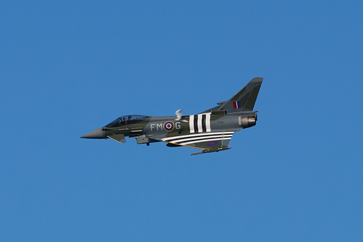 RAF Typhoon fighter jet in flight against clear blue sky, displaying D-Day invasion stripes.