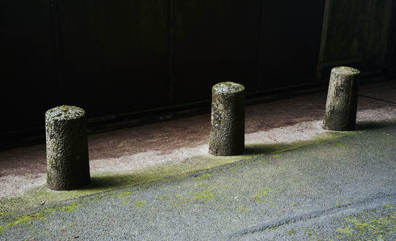 Three weathered stone bollards in a row casting shadows on a textured pavement with moss growth.