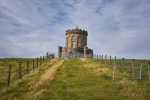 Stone tower with crenellations on a grassy hilltop surrounded by fencing.