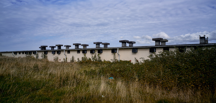 Long white institutional building with multiple chimneys set against overgrown grassland and blue sky.