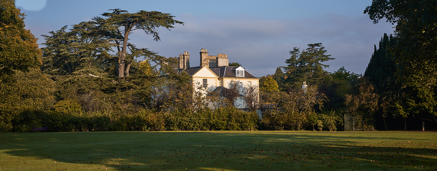 Cream-colored manor house with ornate chimneys surrounded by mature trees and manicured lawn.