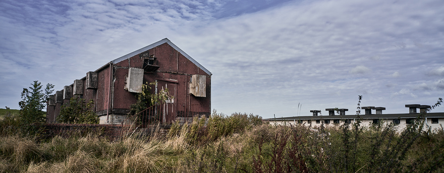Abandoned industrial brick structure with boarded windows amid overgrown vegetation under partly cloudy sky.