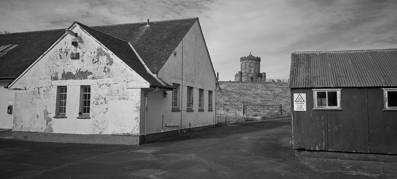 White-washed cottage beside dark buildings with stone tower on hillside in background, black and white photograph.