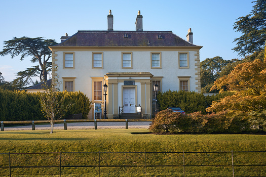 Cream-colored Georgian mansion with dark roof framed by autumn foliage and manicured grounds.