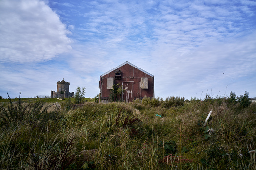 Abandoned red barn structure on overgrown hillside beneath blue sky with clouds.
