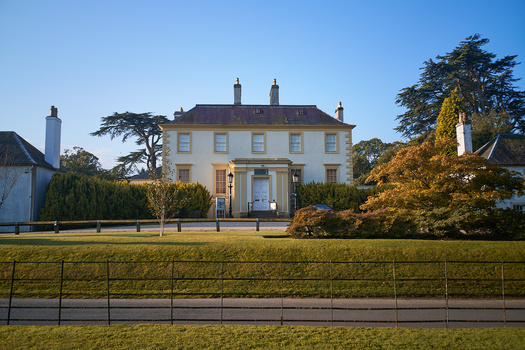 Cream-colored Georgian mansion with dark roof set behind iron fence, photographed in bright daylight with autumn foliage.