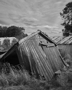 Collapsed corrugated metal structure tilted at steep angle in overgrown field under cloudy sky.
