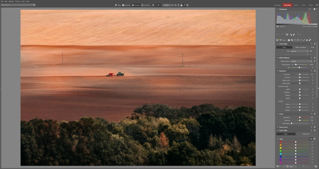 Aerial view of a red tractor working in a vast field at sunset with forested treeline below.