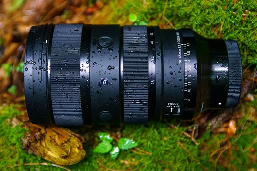 Black telephoto lens with water droplets resting on moss and forest debris.