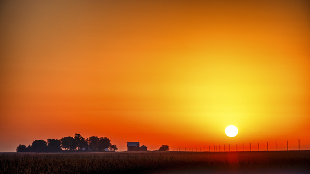 Bright sun on horizon at sunset over rural landscape with silhouetted farm buildings and windmill.
