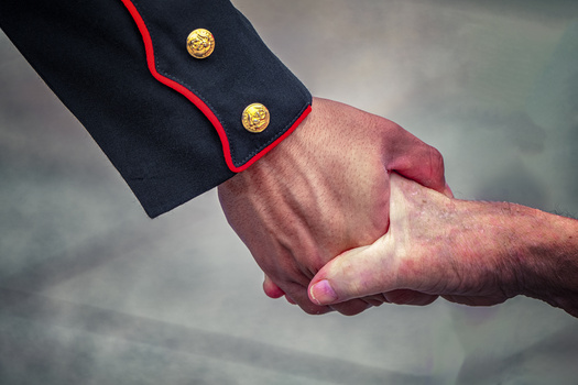 Two hands clasping in a handshake, one wearing a formal navy jacket with red trim and gold buttons.