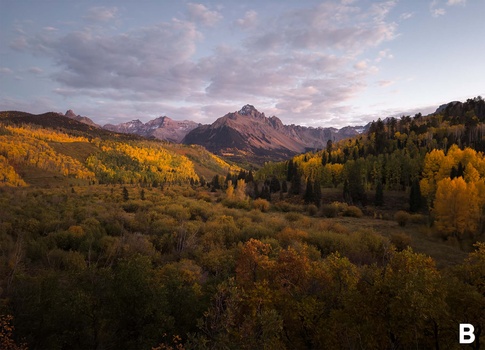 Mountain valley landscape with golden aspen trees in fall, evergreen forests, and snow-capped peaks under dramatic cloudy sky.