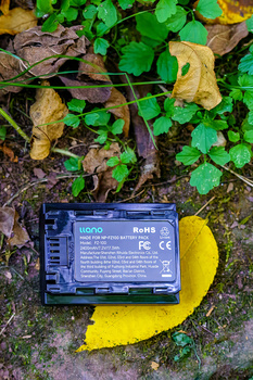 Camera battery placed on yellow leaf surrounded by green foliage and fallen leaves on ground.
