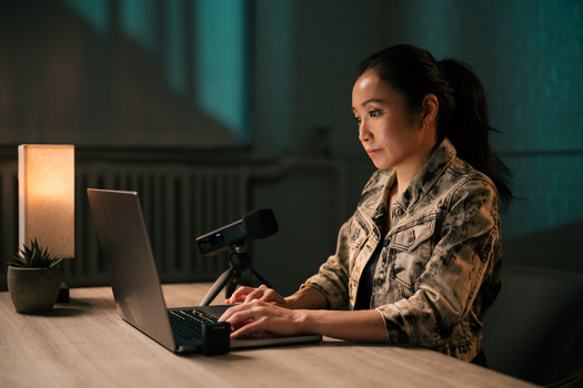 Woman speaking into microphone while typing on laptop at desk with warm lamp lighting.