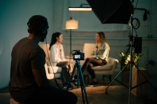 Camera on tripod in foreground of a studio interview setup with two people seated on a couch.