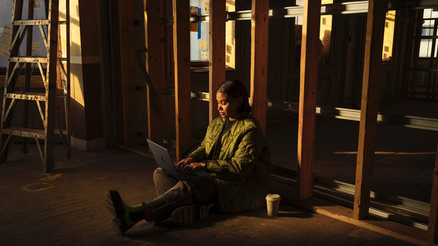 Person sitting on floor working on laptop in dimly lit interior space with wooden columns and warm window light.