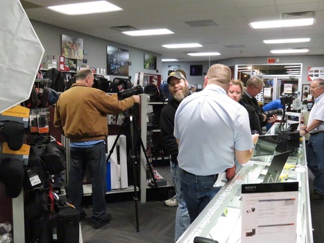 A group of photographers and crew members gathered in a retail electronics store during what appears to be a commercial shoot or event.