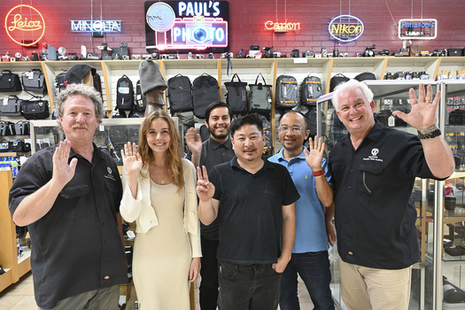 Six people waving at camera inside a photography equipment retail store with neon brand signs.
